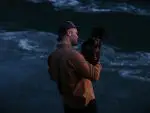 Country singer Zach Bryan stands in front of the ocean holding a dog