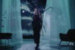 In an article about Kim Kardashian in "American Horror Story Delicate," Kim Kardashian stands in a black dress with a white wig on in front of draping white curtains in blue-ish lighting.