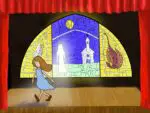 A young girl in a blue dress walks off a stage framed by a red curtain. A stained glass window hangs in front of a black background, depicting a church in a field against a sea-blue sky and a young woman glowing with divinely white light. Two images flank it, an angel to the left and a dragon to the right, framed in yellow.